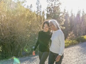 A beautiful mixed race young adult woman embraces her vibrant retirement age mother. The mother and daughter are enjoying a relaxing walk in nature on a beautiful, sunny day. In the background is a mountainous evergreen forest bathed in sunlight.