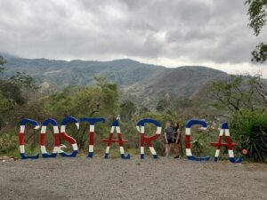 Two Sanford Health doctors stand in place of the "i" in a sign spelling out "Costa Rica" with a beautiful view of mountains.