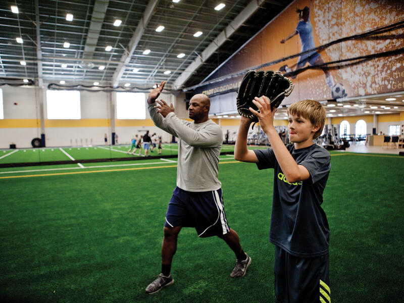 Sanford Sports Academy baseball instructor models a catching position for a student next to him on fieldhouse turf.