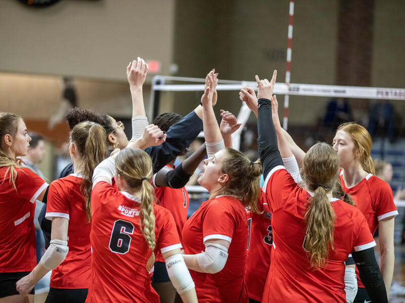 A volleyball team in red uniforms celebrates a win with their arms in the air.