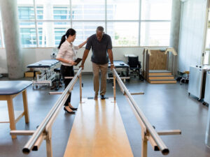 Female physical therapist helps a senior man walk following a stroke. The man is using parallel bars in a rehab center.