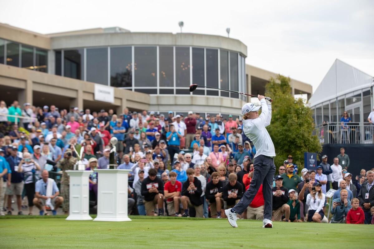 Teen boy follows through on a golf swing in front of a crowd of spectators.