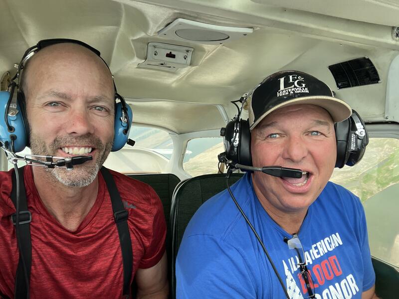 Two smiling men wear headsets inside a small airplane high above the Missouri River.