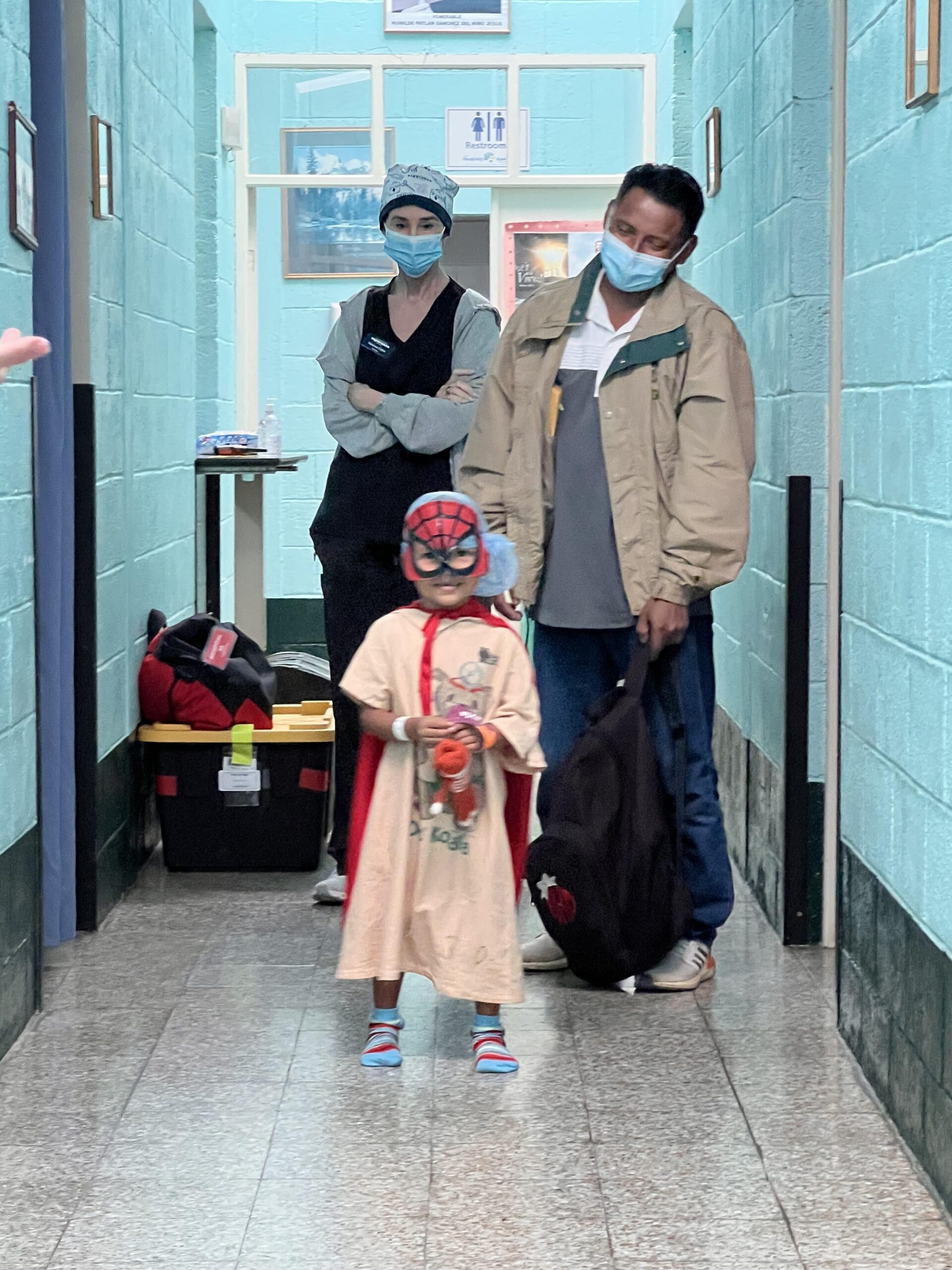 A young patient in surgical scrubs and a Spider-Man mask poses with his father and a Sanford Health nurse in the hallway of a Guatemalan hospital.