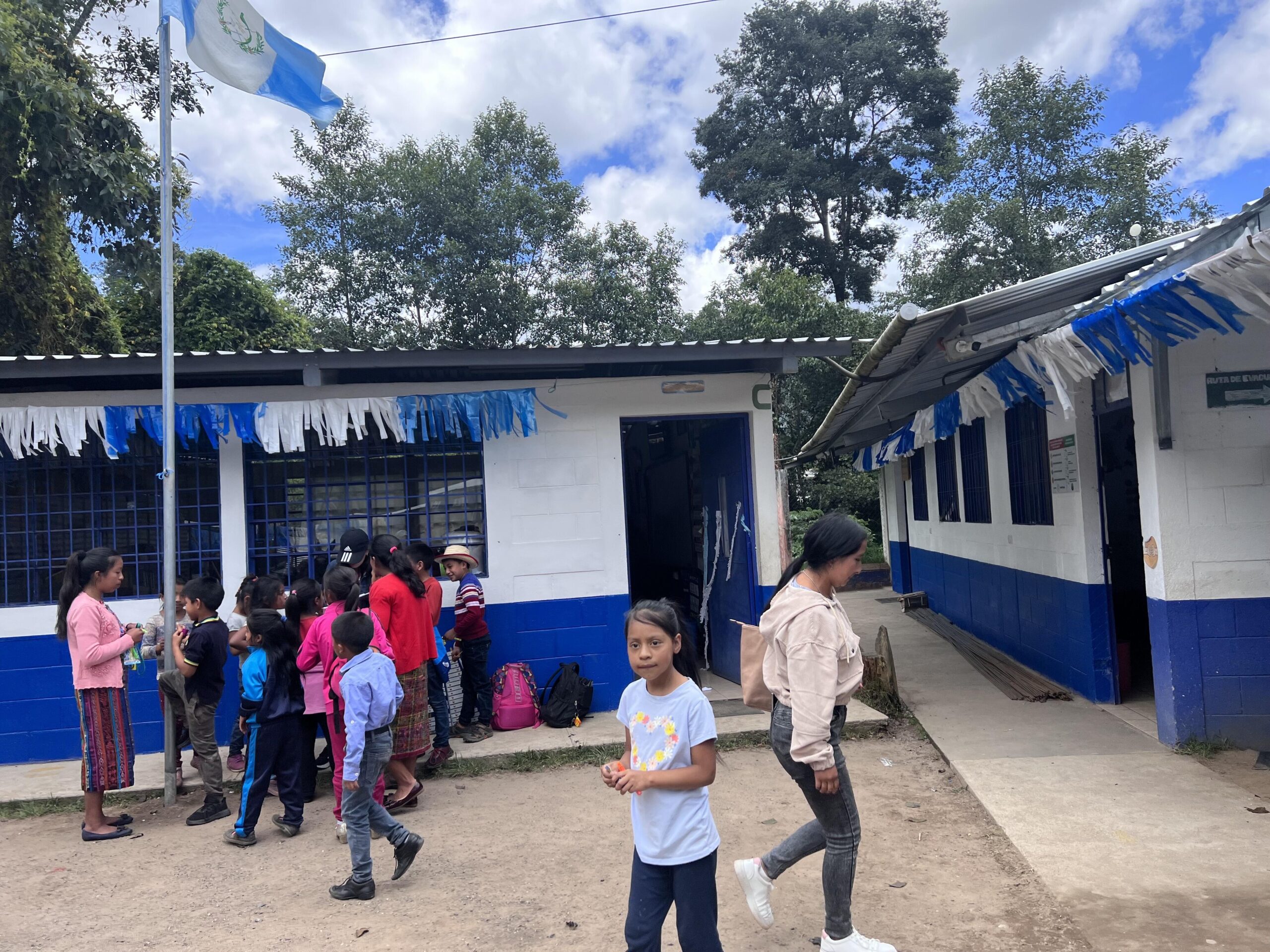 Children and teachers in a dirt courtyard between cinderblock buildings with blue and white banners.