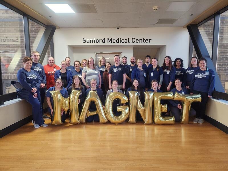 Nurses and nursing leaders in matching blue shirts pose with shiny balloons that spell "MAGNET" under a sign for Sanford Medical Center.