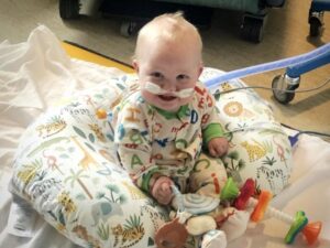 Smiling baby hospital patient sits playing while oxygen tubes are in his nose.