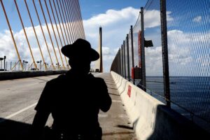 A silhouette of a law enforcement officer walking along a sunny bridge lined with suicide-prevention barriers.