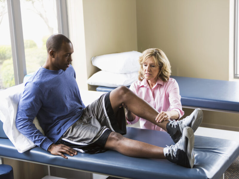 A female physical therapist examining a young Black man on a treatment table. He is sitting on the table with his legs stretched out in front of him. The physical therapist, a senior woman, is examining his knee. They are both looking at his bent knee with serious expressions on their faces.