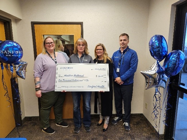 Four people stand holding a giant check made out to Heather Malterud. Blue balloons decorate the hallway.