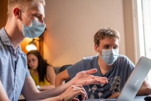 Two students in surgical masks gestures while sharing a laptop screen.