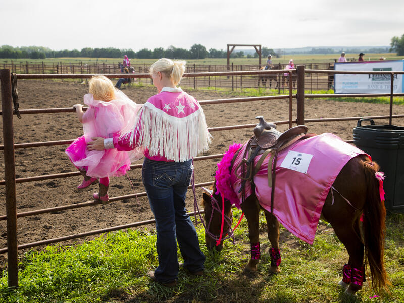 Two cowgirls dressed in pink fringe watch other rodeo competitiors while their horse grazes.