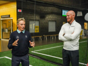 Mature golfer talks with smiling trainer on an indoor field.