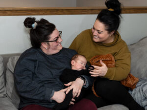 Two moms hod their infant son who has a breathing tube in his nose.