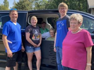 Two mature adults sit in a van while four family members stand outside.