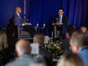 Two businessmen sit on stage in front of a crowd.