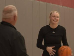 College basketball player laughs with coach at practice inside gym.