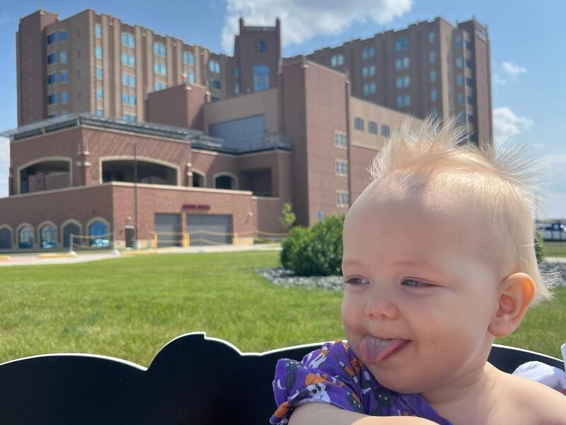 Playful 9-month-old boy sticks his tongue out while riding in a wagon outside a hospital on a sunny day.