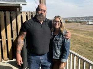 A man stands with his arm around his wife on their back porch. He's serious; she's smiling.