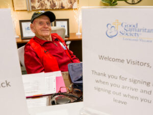Senior man softly smiles from a desk between two welcome signs.
