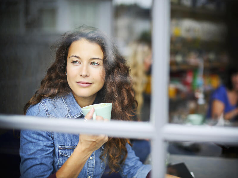 Lifestyle image of woman looking out of a cafe window whilst holding a drink.