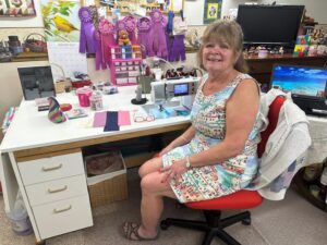 Older adult woman sits at her sewing table with blue and purple competitive ribbons on display above it.
