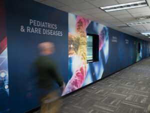 Wall mural with microscope images of genetic material between labels including "Pediatrics & Rare Disease." A person is walking by in a blur.