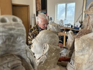 Stone sculptures of people frame a senior man using a chisel in his studio.