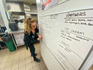 Woman writes on whiteboard in kitchen, with categories including vegetables, ice cream, cookies and desserts.