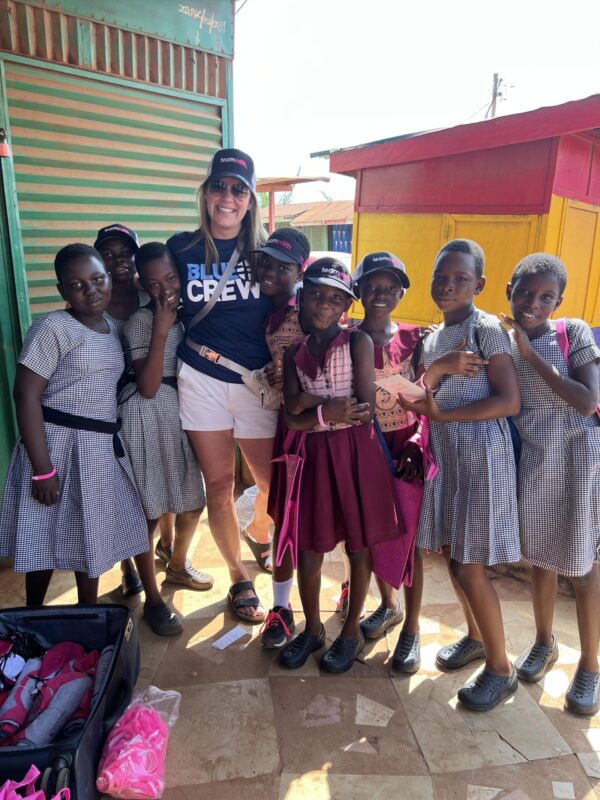Tall blonde woman smiles with a group of Ghana schoolgirls in uniform dresses.