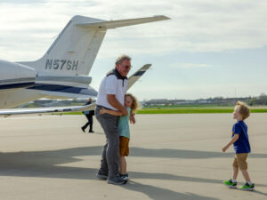 Mature man happily hugs one child while reaching for another near the tail of a small airplane.