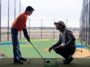 14-yearold boy stands with a golf club while a man kneels to talk to him above a driving range.