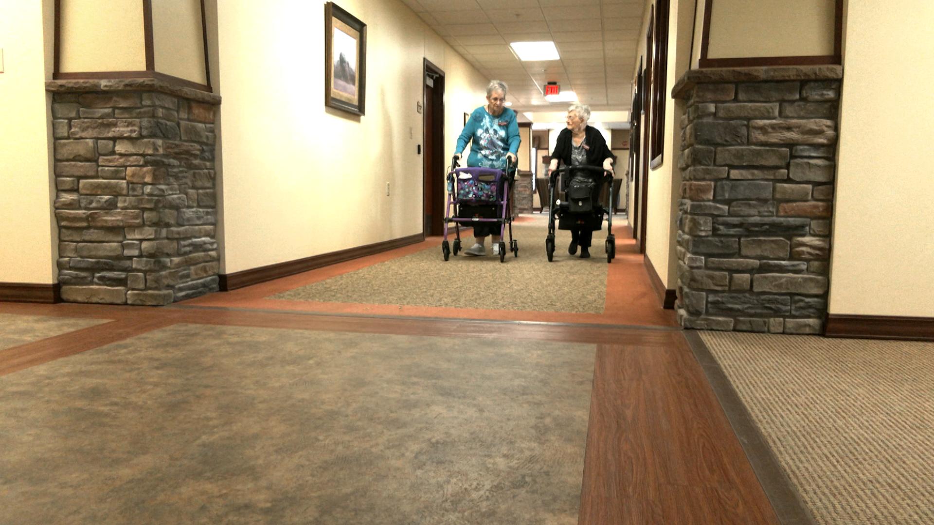 Two senior women walk down a hallway using walkers.