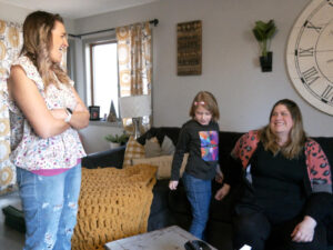 Two women laugh in a living room with a 7-year-old girl playing between them.