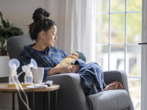 A new mother sitting on a rocking chair in her living room wearing a robe holding her newborn baby. There is a breast pump and nursing supplies around.