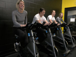 Four friends laugh while riding stationary bikes in a gym.