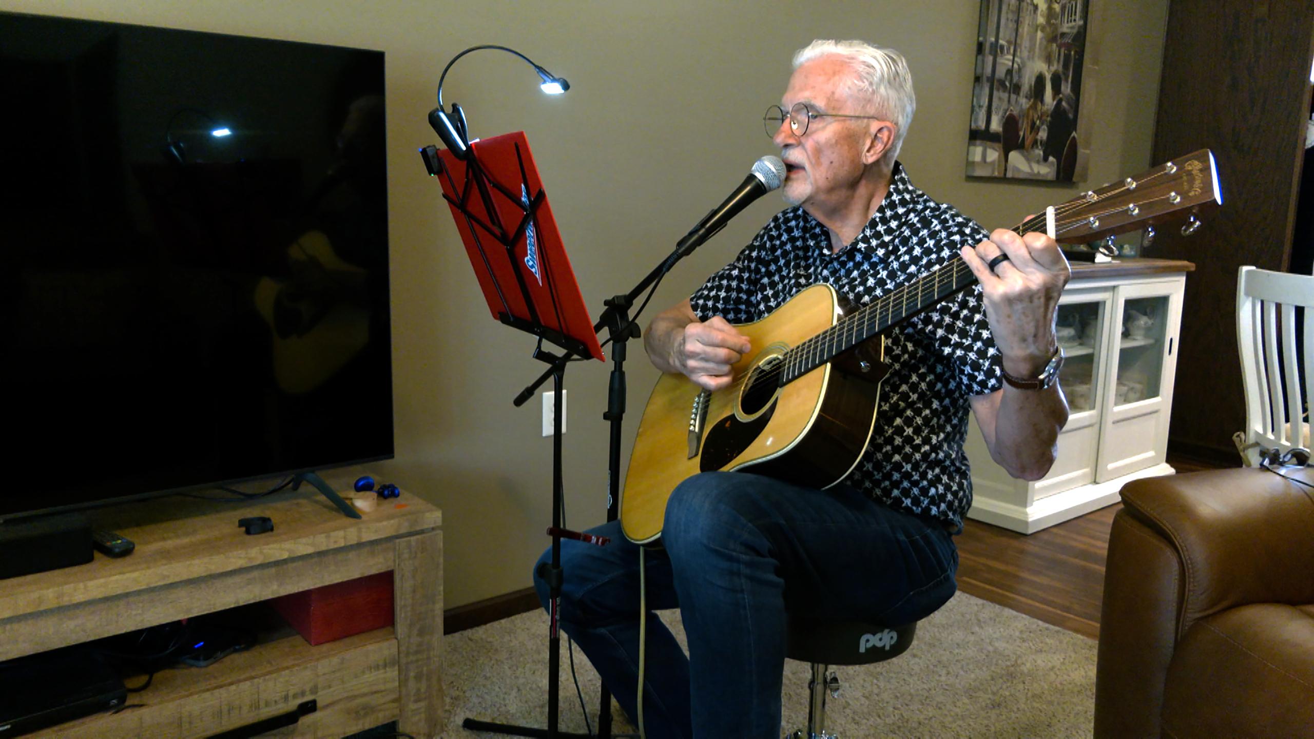 Senior man plays guitar and sings into a microphone at home.