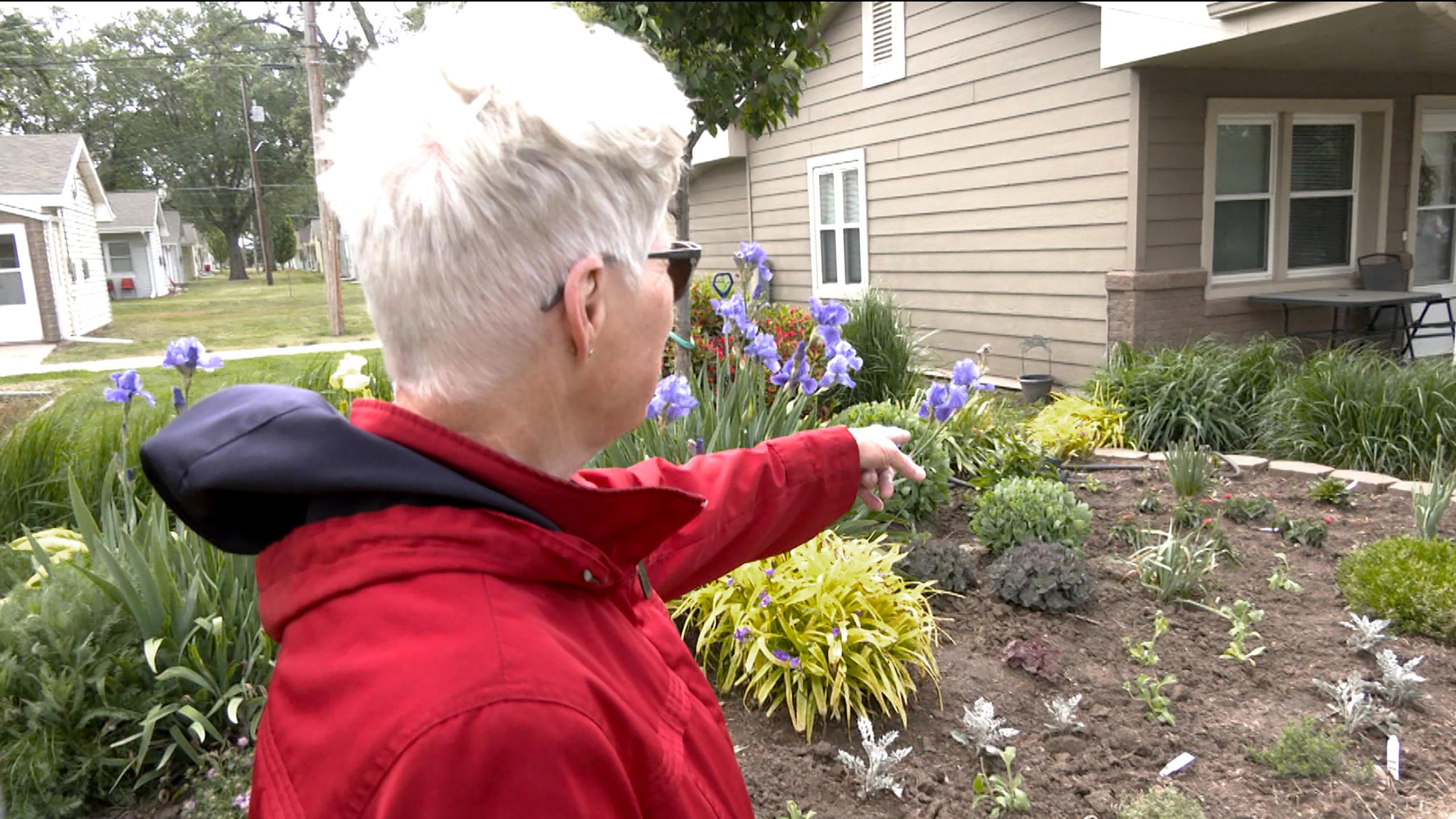Senior woman points to flowers in a new garden bed beside a house.