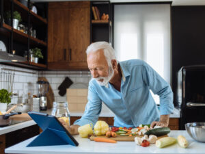 A senior man watching a tablet while busy making healthy food in the kitchen.