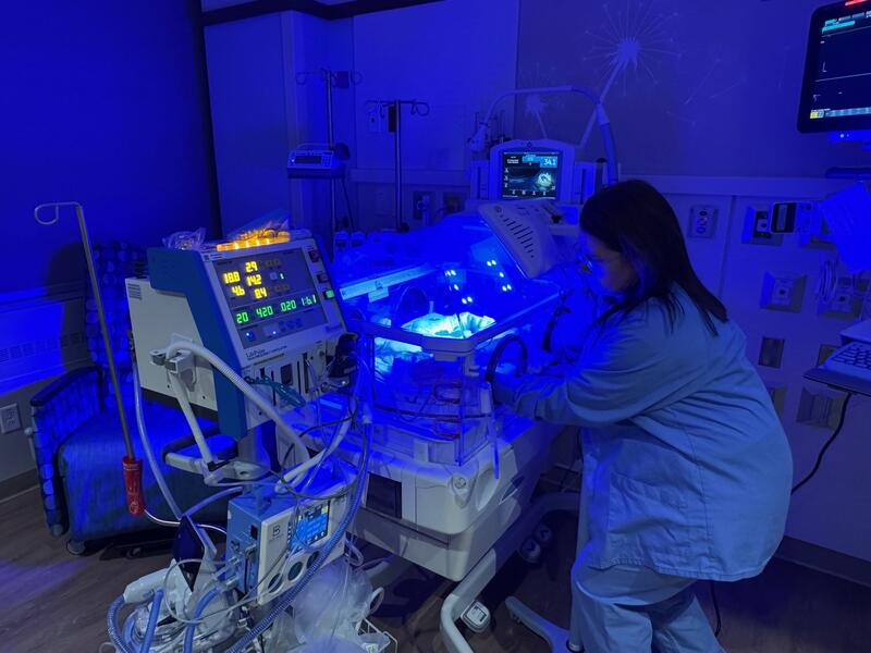 Nurse works in a hospital nursery under blue lights and computer monitors.