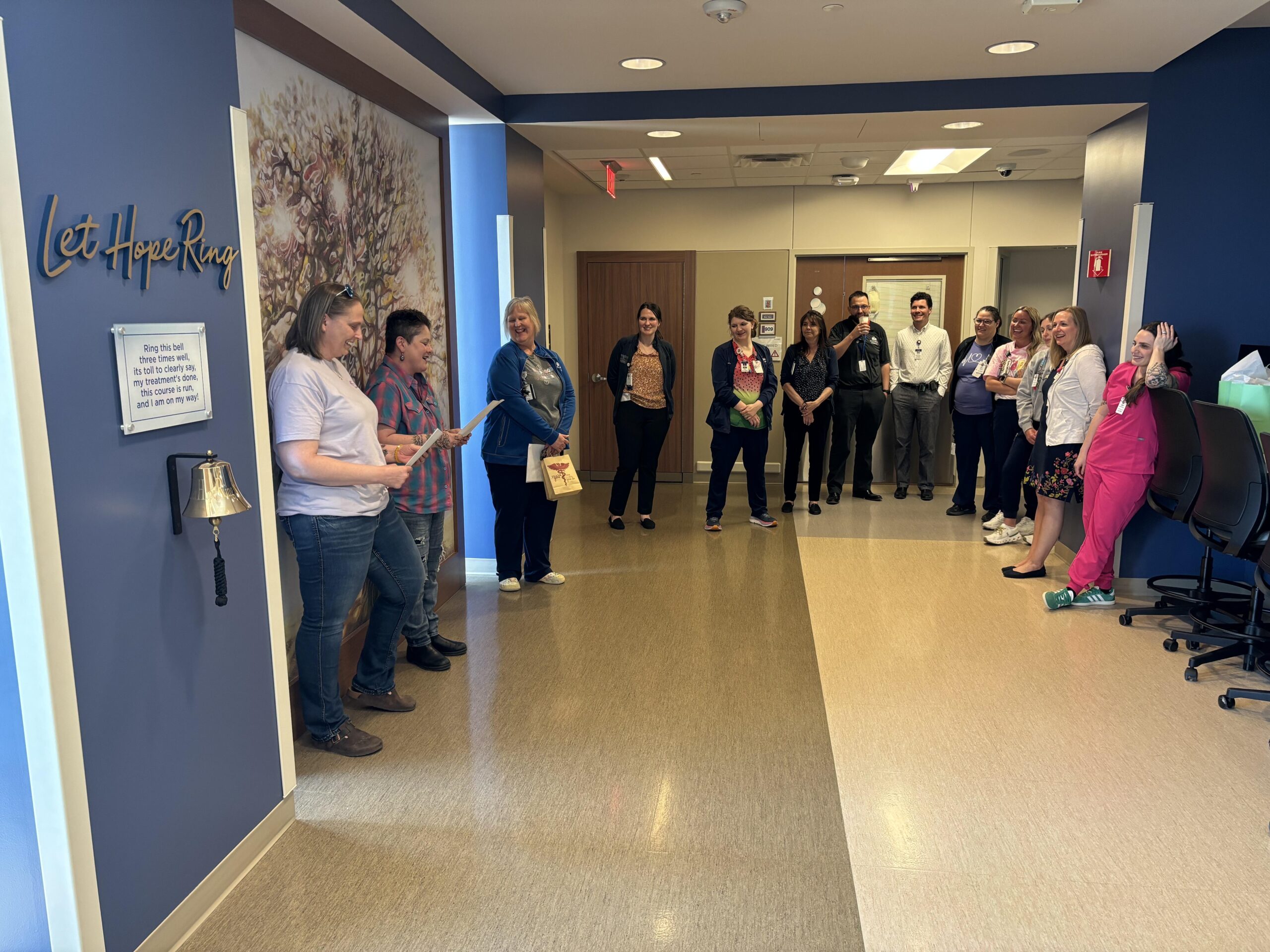 Two happy women read letters to a semicircle of nurses and other hospital workers standing and smiling in a hospital hallway.