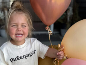 Excited 3-year-old girl holds pink and gold balloons and wears a t-shirt that says "threenager."