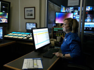 Businesswoman sits in darkened control room with computer and television screens lit up.