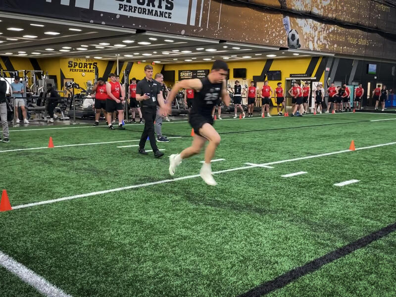 A high school student runs across a football field while trainers and other athletes watch.
