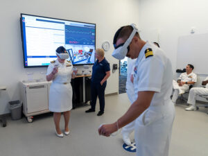 Woman and man in U.S. Navy dress white uniforms test VR headsets as others watch their work on a large wall-mounted screen.