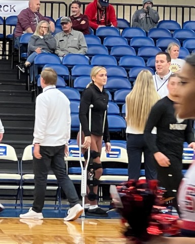 Girl on crutches stands at sidelines of a basketball court while fans, athletes and coaches blur around her.