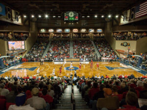 Wide view of vintage style basketball arena with digital and analog timers, men's college players running across parquet flooring and fans filling the stands.