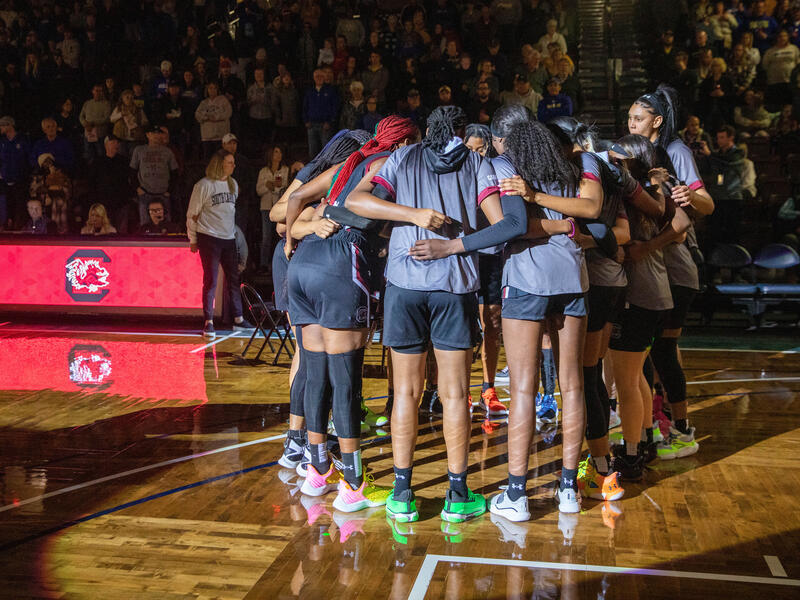 Women's college basketball players huddle on a parquet basketball floor in front of a packed crowd.