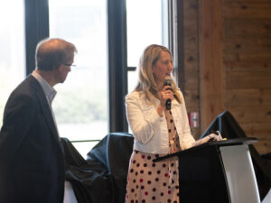 Businesswoman speaks into a microphone at a podium with a businessman turning to listen nearby.