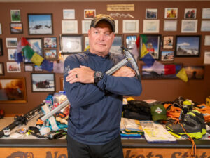 Midlife man crosses arms and carries an icepick in a room decorated with travel photos and mountain-climbing gear.